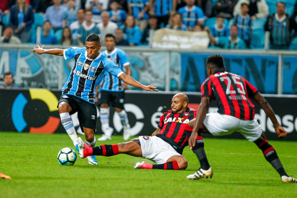 atleta do Atlético Goianiense aplica carrino para recuperar bola enquanto jogador do Grêmio tenta driblar com a perna direita