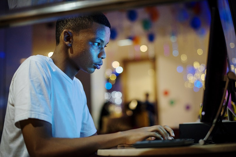 Um jovem está sentado com as mãos sobre um teclado de computador, seu rosto está iluminado pela tela.